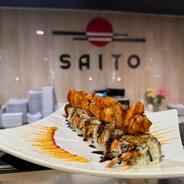 A sushi roll with shrimp tempura and sauce on a white plate, with the Saito logo in the background.
