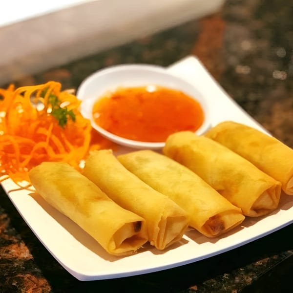 Five golden-brown spring rolls with shredded carrots and sweet chili sauce on a white plate.