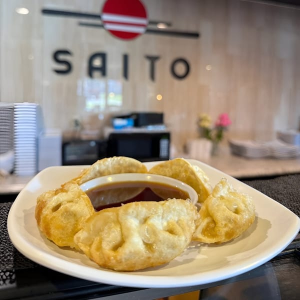Crispy fried dumplings with dipping sauce on a white plate, with the Saito Japanese Steakhouse sign in the background.
